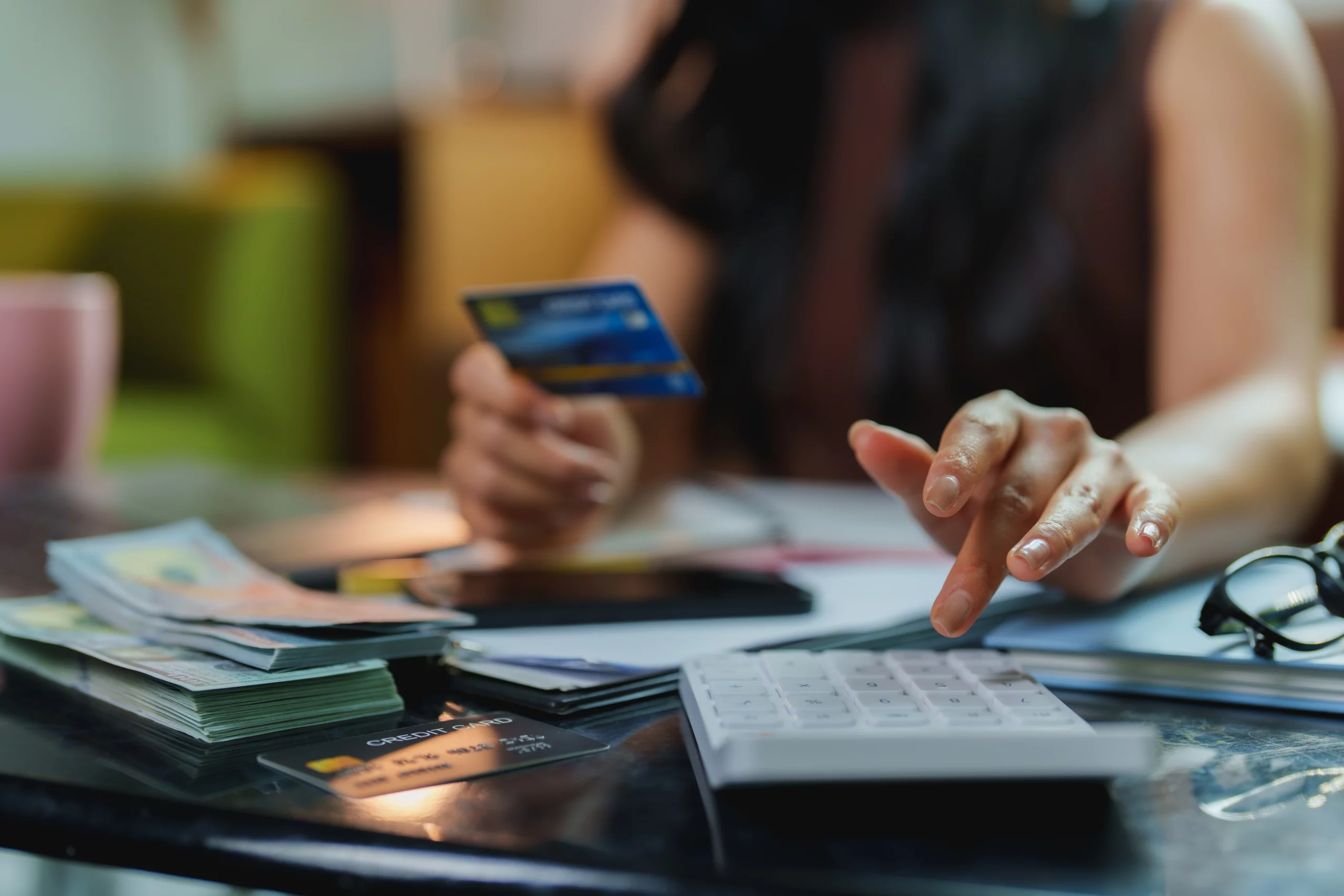Person using calculator and holding credit card on table with banknotes, smartphone, and glasses, illustrating credit card debt responsibility and financial planning related to authorized user credit card issues.