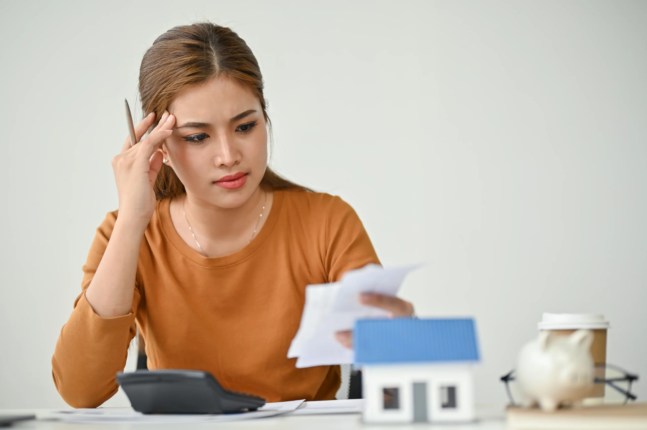 Concerned woman reviewing financial papers at a table with a calculator, piggy bank, and house model, illustrating stress over homeowners association (HOA) fees and how they may still be owed even after filing bankruptcy.